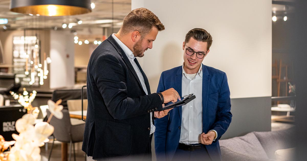 Zu sehen sind zwei Verkäufer im Ausstellungsraum. Beide tragen einen Anzug mit weißem Hemd. Der Verkäufer links ist seitlich zu sehen und hält ein Tablet, auf das er dem anderen Verkäufer etwas zeigt.