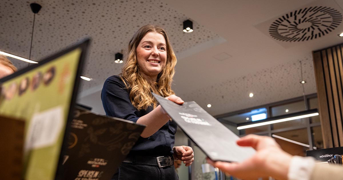 Eine junge Frau mit welligem, braunem Haar und dunkler Arbeitskleidung reicht eine Menükarte an eine Person mit heller Kleidung. Im Vordergrund sind weitere Menükarten sichtbar. Der Raum ist modern gestaltet mit Holzpaneelen, Deckenleuchten und Akustikplatten.