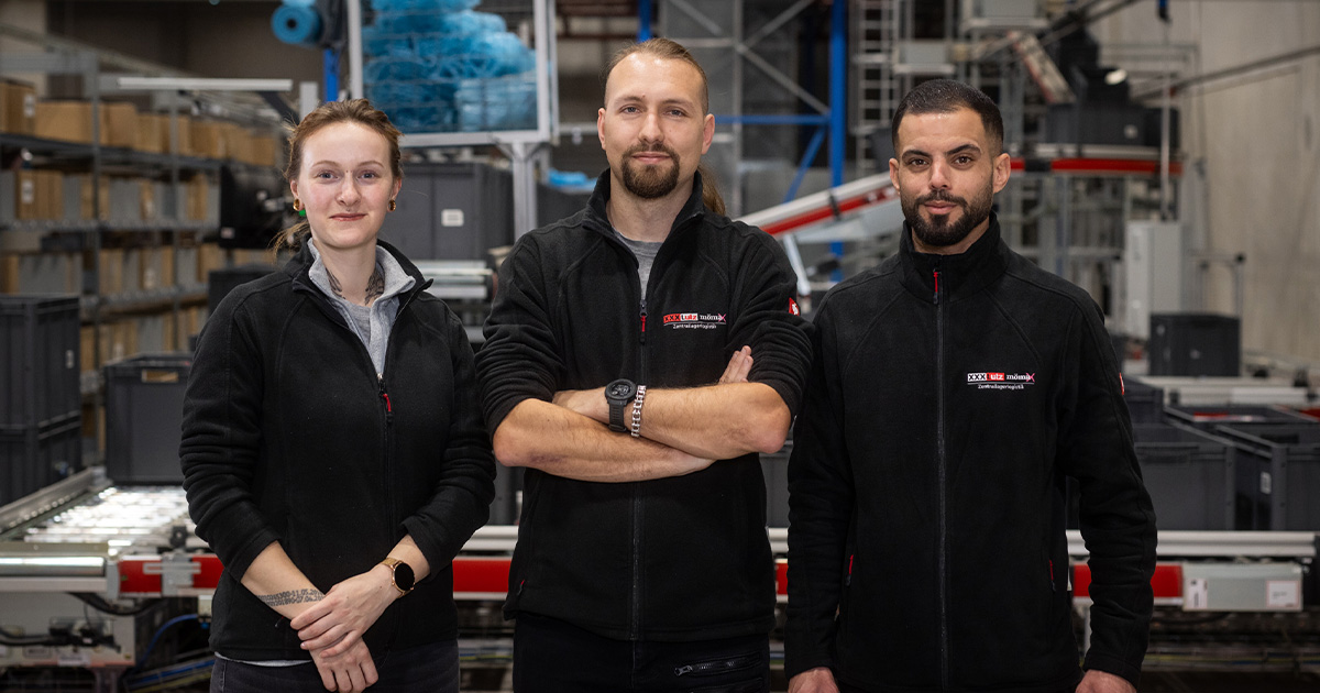 Drei Mitarbeitende in schwarzer Arbeitskleidung stehen nebeneinander in einer Lagerhalle. Eine Frau und zwei Männer blicken frontal in die Kamera. Im Hintergrund sind Förderanlagen, Regale und industrielle Strukturen zu sehen.