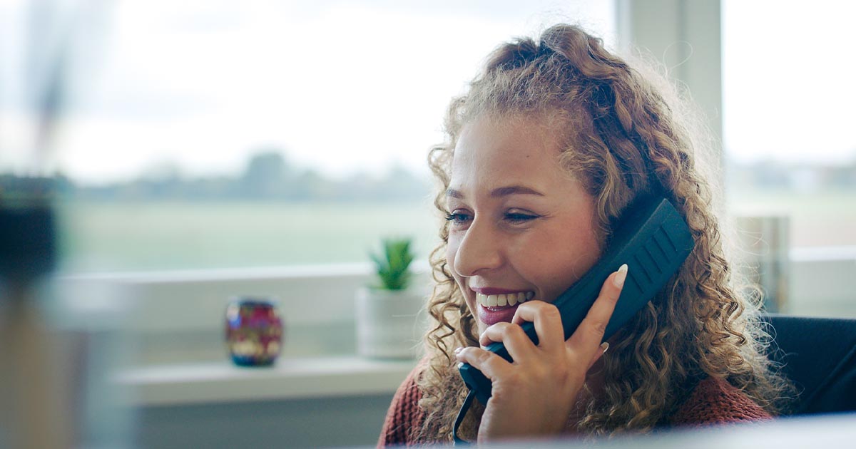 Eine weibliche Mitarbeiterin sitzt an ihrem Schreibtisch im Büro und telefoniert. Sie hält den Telefonhörer ans Ohr und lächelt. Im Hintergrund ist ein Fenster mit Blick auf eine grüne Landschaft sowie Dekoration auf der Fensterbank zu sehen.