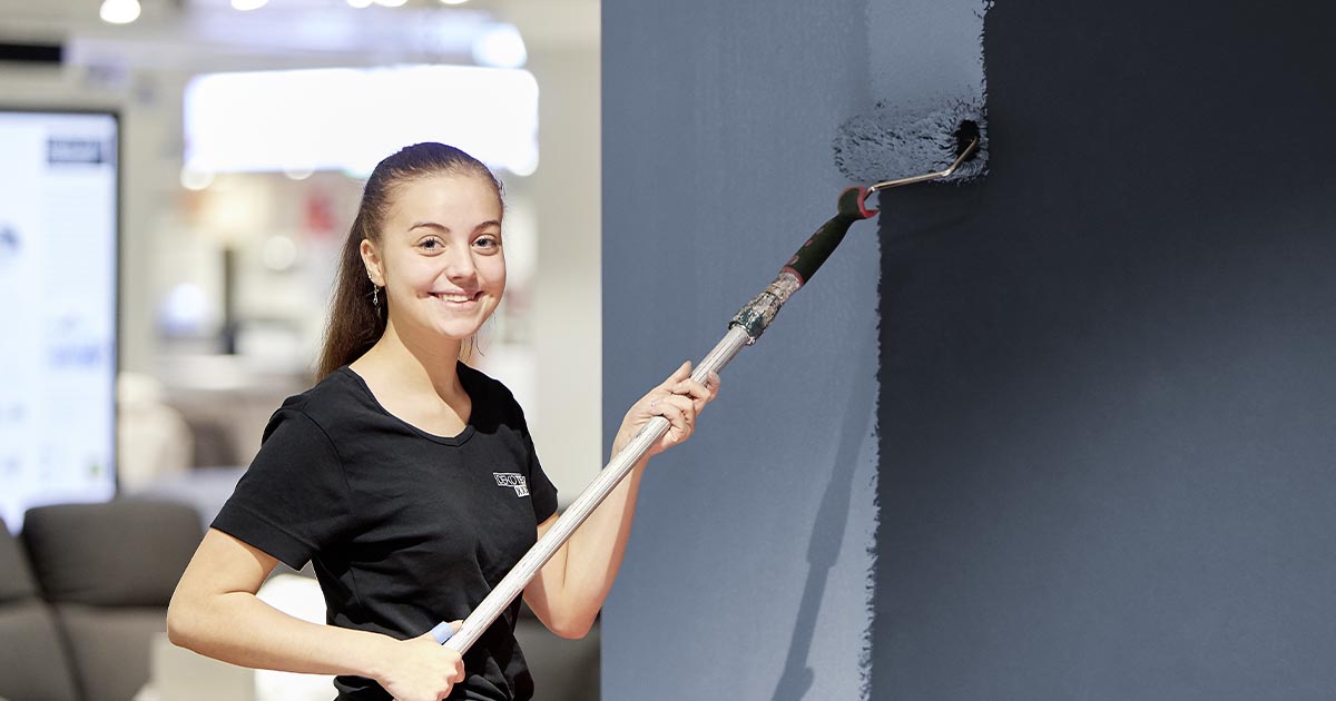 Eine junge Frau in schwarzer Arbeitskleidung streicht mit einer Farbrolle eine Wand dunkelblau. Sie lächelt in die Kamera. Im Hintergrund ist ein Verkaufsraum mit unscharfen Möbeln sichtbar.