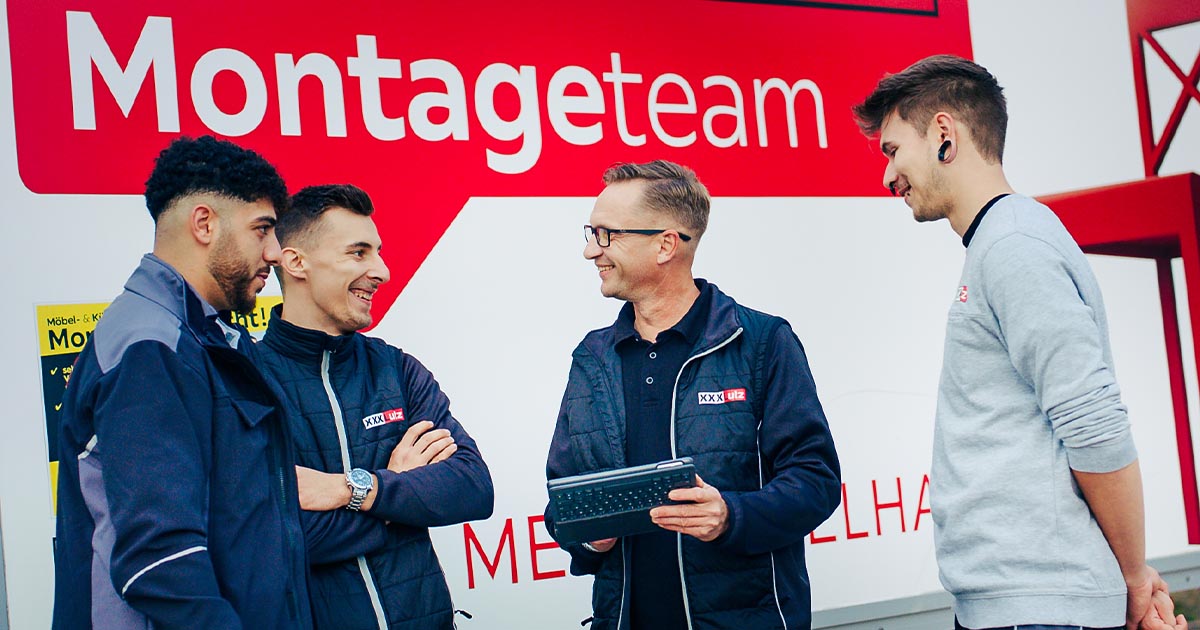 Vier Mitglieder des Montageteams von XXXLutz stehen vor einem Montage-LKW auf dem "Montageteam" steht und das Markenzeichen der "rote Stuhl" abgebildet ist. Sie unterhalten sich lächelnd. Einer der Männer hält ein Tablet, während die anderen in Arbeitskleidung stehen und zuhören. 