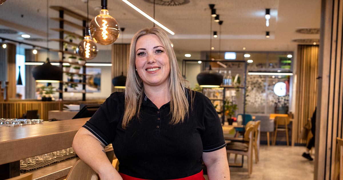 Eine blonde Frau in schwarzer Arbeitskleidung mit roter Schürze lehnt lächelnd an einer Theke in einem modern eingerichteten Restaurant. Im Hintergrund sind hängende Lampen, Pflanzen und Sitzmöbel zu sehen.