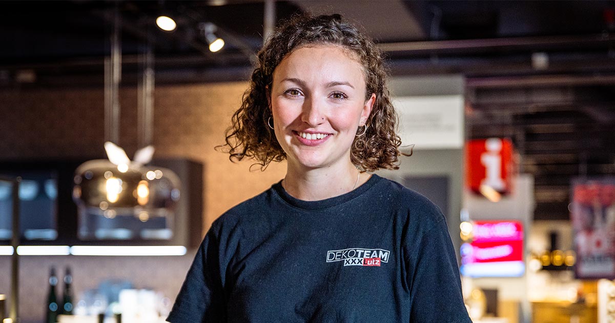 Eine junge Frau mit lockigem Haar steht lächelnd in einem modern gestalteten Küchenbereich. Sie trägt ein schwarzes T-Shirt mit dem Aufdruck "Dekoteam XXXLutz". Im Hintergrund sind dekorative Hängelampen, Küchenmöbel und leicht verschwommene, beleuchtete Bereiche zu erkennen.
