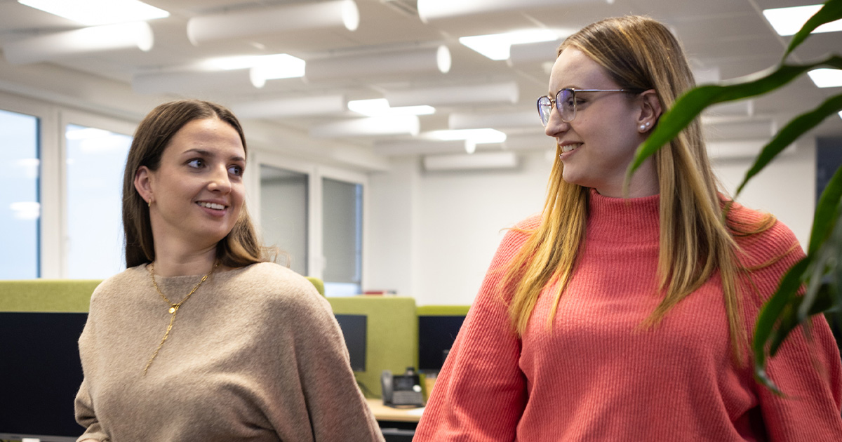 Zwei Frauen gehen nebeneinander durch ein Büro und unterhalten sich. Die Frau links trägt einen beigefarbenen Pullover und eine schwarze Hose, die Frau rechts trägt ein korallfarbenes Oberteil und eine dunkle Hose. Im Hintergrund sind Schreibtische, Monitore und eine helle Büroeinrichtung zu sehen.