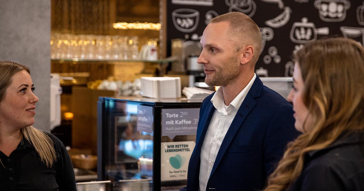 Ein Mann in einem dunkelblauen Anzug spricht mit zwei Frauen in Arbeitskleidung in einem Café- oder Restaurantbereich. Im Hintergrund sind gläserne Regale, eine schwarze Wand mit weißen Symbolen und ein Kühlbereich mit Lebensmittelauslagen zu sehen.