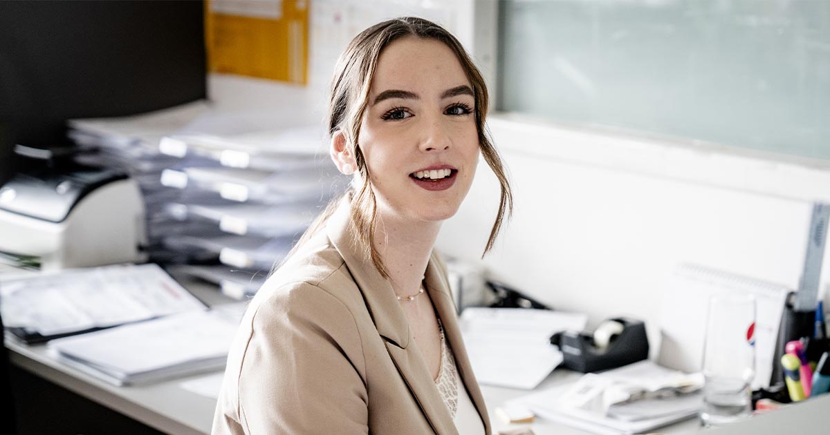 Eine weibliche Mitarbeiterin sitzt an ihrem Schreibtisch im Büro und schaut lächelnd in die Kamera. Sie trägt ein helles Blazer-Outfit. Im Hintergrund sind Aktenstapel, ein Drucker und eine Glastafel mit Notizen zu sehen.