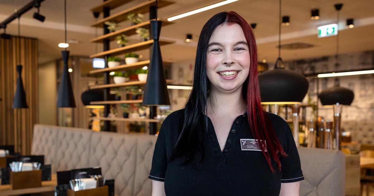 Eine junge Frau mit schwarz-rotem Haar und schwarzer Arbeitskleidung steht lächelnd in einem modern eingerichteten Restaurant. Im Hintergrund sind gepolsterte Sitzmöbel, hängende Lampen und Regale mit Pflanzen zu sehen.