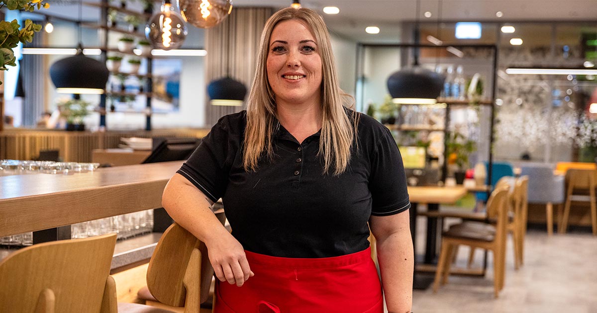 Eine Frau in einem modernen Restaurantumfeld steht an einer hölzernen Bartheke und lächelt in die Kamera. Sie trägt ein schwarzes Polohemd und eine rote Schürze. Im Hintergrund sind stilvolle Hängelampen, Holzmöbel und Pflanzen zu sehen.