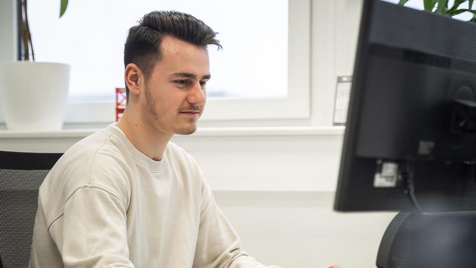Ein junger Mitarbeiter mit dunklen Haaren arbeitet konzentriert an einem PC-Arbeitsplatz im Büro. Im Hintergrund steht eine große Pflanze.