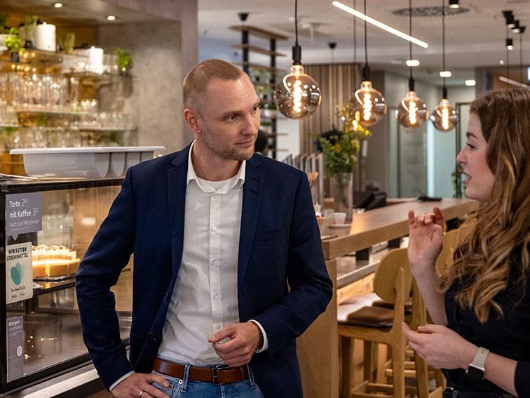 Ein Mann in einem blauen Anzug und Jeans steht mit drei Frauen in schwarzer Arbeitskleidung in einem modern eingerichteten Restaurantbereich. Sie unterhalten sich, die Frau ganz rechts gestikuliert mit ihren Händen. Im Hintergrund sind eine gläserne Theke mit Torten, Regale mit Gläsern, hängende Lampen und Holzmöbel zu sehen.