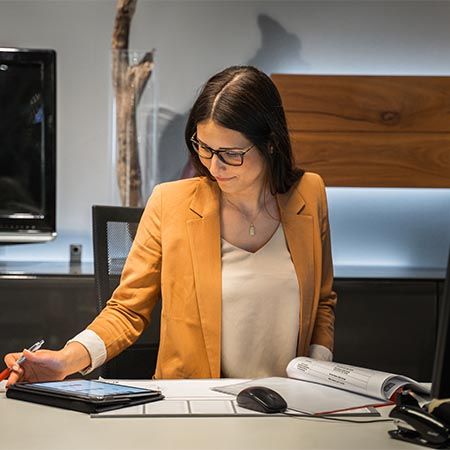Eine Frau mit dunklem, schulterlangem Haar und Brille sitzt an einem Schreibtisch. Sie trägt einen orangefarbenen Blazer über einer cremefarbenen Bluse. Mit einem Stift in der Hand arbeitet sie an einem Tablet, während sie gleichzeitig auf Unterlagen blickt. Im Hintergrund sind moderne Einrichtungsgegenstände und eine Holzverkleidung zu sehen.