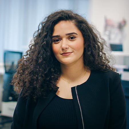 Eine Frau mit langen, lockigen Haaren steht in einem Büro und blickt mit einem dezenten Lächeln in die Kamera. Sie trägt eine schwarze Jacke über einem schwarzen Oberteil. Im Hintergrund sind unscharf ein Schreibtisch, ein Monitor und weitere Büroeinrichtung zu erkennen.