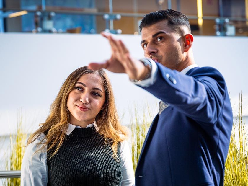 Führungskraft und Azubine stehen im Verkaufsraum. Beide sind chic gekleidet und frontal zu sehen. Er hält seinen Arm ausgestreckt auf Schulterhöhe als würde er auf etwas in der Ferne zeigen. Die Azubine schaut lächelnd in die Ferne in die gezeigte Richtung.
