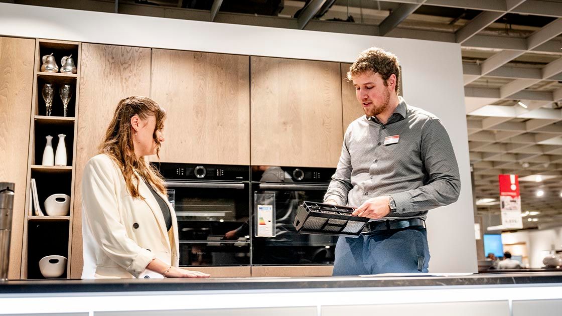 Zu sehen sind zwei Verkaufsmitarbeiter vor einer Ausstellungsküche aus Holz. Der Verkäufer rechts hat ein Filtersystem eines Boras-Abzugssystem in der Hand und erklärt der Azubine links die Funktionalität