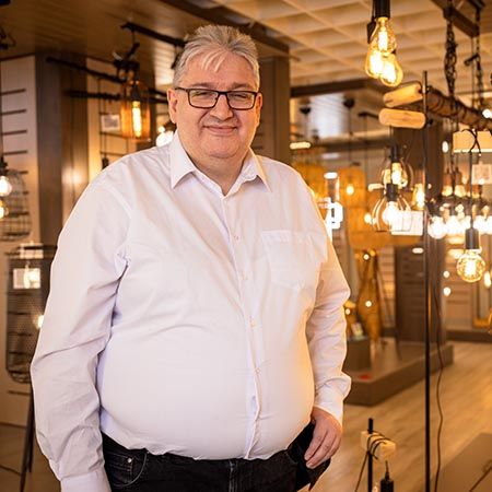 Zu sehen ist ein Verkäufer in der Leuchten-Ausstellung eines Verkaufshauses. Er steht frontal zur Kamera und lächelt. Er trägt ein weißes Hemd und eine schwarze Brille. Die Lampen um in herum sind angeschaltet und leuchten weiß-gelblich. 