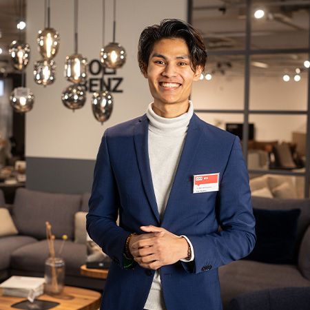 Ein junger Mann in einem blauen Anzug und einem weißen Rollkragenpullover steht lächelnd in einer modern eingerichteten Möbelabteilung. Im Hintergrund sind Sofas, dekorative Beleuchtung und weitere Möbelstücke zu erkennen.