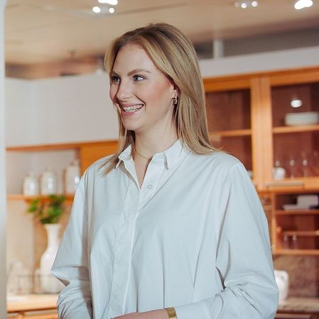 Eine Frau in einem weißen Hemd steht lächelnd zu einer Person außerhalb des Bildes in einem stilvoll eingerichteten Bereich eines Möbelhauses. Im Hintergrund sind Regale mit Dekorationen und warmes Licht zu sehen.