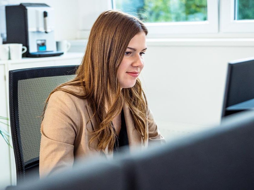 Die Frau in einem beigen Blazer sitzt an einem Schreibtisch und arbeitet konzentriert. Im Hintergrund sind eine Kaffeemaschine und ein Fenster mit Blick ins Grüne sichtbar. Die Szene vermittelt eine moderne und professionelle Arbeitsumgebung.