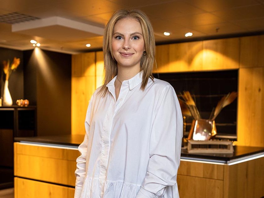 Zu sehen ein Portrait einer jungen Frau in einer weißen Bluse. Sie steht vor einer Ausstellungsküche aus Holz, die von Deckenspots angeleuchtet werden.