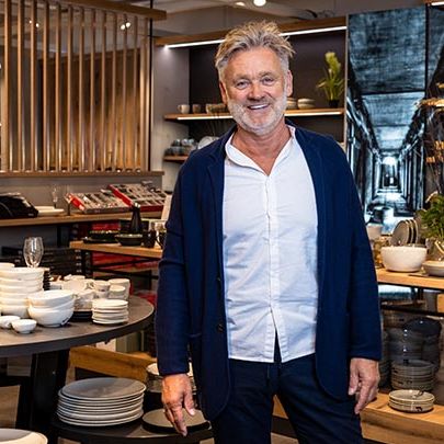 Der Einkaufsmitarbeiter mit grauem Haar steht im weißem Hemd lachend frontal zur Kamera. Im Hintergrund sieht man die Ausstellungstische und Wandregale voller Geschirr, wie Teller, Gläser, Besteck in weiß, dunkelgrün, beige. 