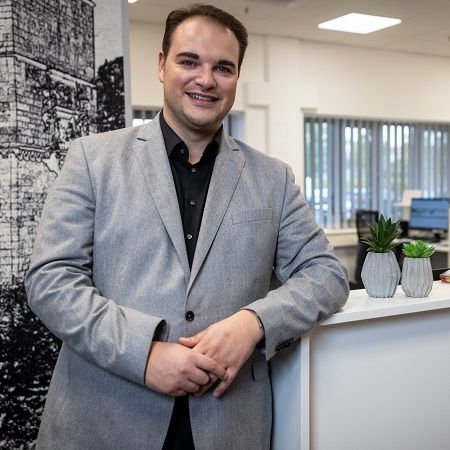 Ein männlicher Mitarbeiter steht in einem modernen Büro an einer weißen Theke und lächelt in die Kamera. Er trägt ein graues Sakko über einem schwarzen Hemd. Im Hintergrund sind Schreibtische, Computer und große Fenster mit Jalousien zu sehen.