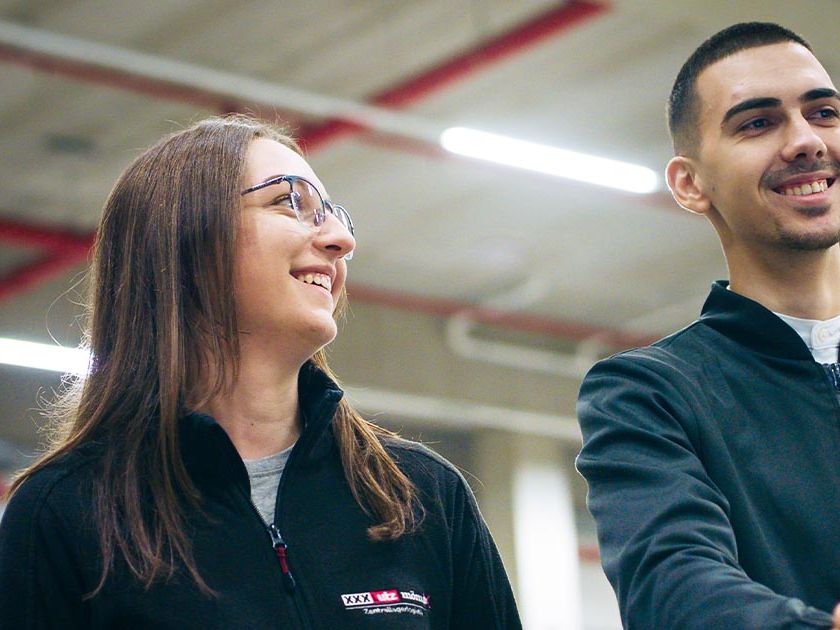 Zwei junge Mitarbeiter in schwarzer Arbeitskleidung stehen lächelnd in einer Lagerhalle. Die Frau trägt eine Brille, und beide wirken entspannt und fröhlich. Im Hintergrund sind rote Rohrleitungen und die Decke der Halle sichtbar.