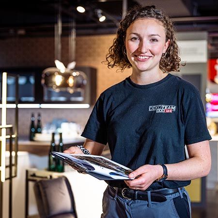 Eine junge Frau mit lockigem Haar steht lächelnd in einem modern gestalteten Küchenbereich und hält ein Klemmbrett. Sie trägt ein schwarzes T-Shirt mit dem Aufdruck "Dekoteam XXXLutz". Im Hintergrund sind dekorative Hängelampen, Küchenmöbel und leicht verschwommene, beleuchtete Bereiche zu erkennen.