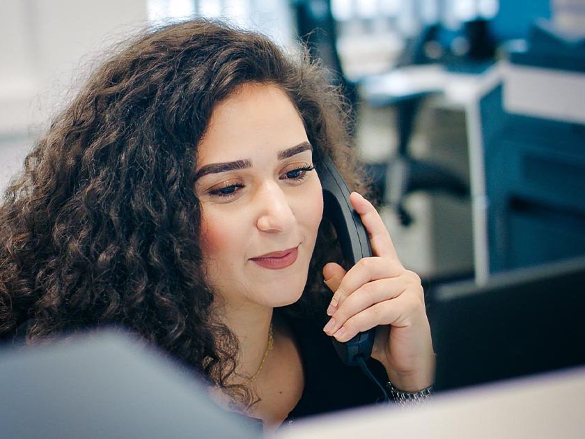 Eine weibliche Mitarbeiterin sitzt an einem Schreibtisch im Büro und hält einen Telefonhörer ans Ohr. Sie hat lockige dunkle Haare und lächelt leicht. Im Hintergrund sind weitere Arbeitsplätze mit Bildschirmen zu sehen.