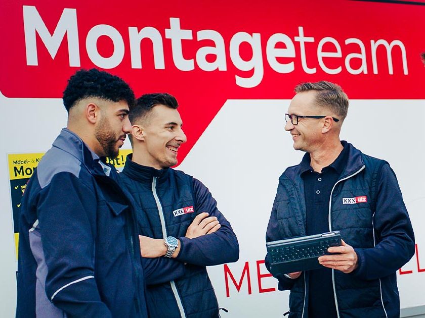 Vier Mitglieder des Montageteams von XXXLutz stehen vor einem Montage-LKW auf dem "Montageteam" steht und das Markenzeichen der "rote Stuhl" abgebildet ist. Sie unterhalten sich lächelnd. Einer der Männer hält ein Tablet, während die anderen in Arbeitskleidung stehen und zuhören. 
