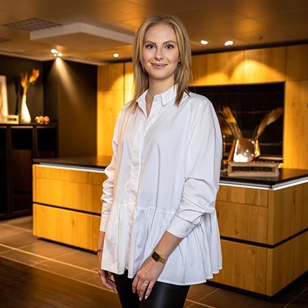 Zu sehen ein Portrait einer jungen Frau in einer weißen Bluse. Sie steht vor einer Ausstellungsküche aus Holz, die von Deckenspots angeleuchtet werden.