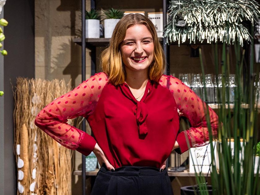Auszubildende zur Einzelhandelskauffrau steht mit Ausstellungsraum für Wohnaccessoires. Sie trägt eine knallrote Bluse und stützt ihre Arme seitlich in der Hüfte ab. Sie lacht herzhaft mit geschlossenen Augen. Um sie herum stehen Orchideen, Gläser und hängt silbernes Lametta.