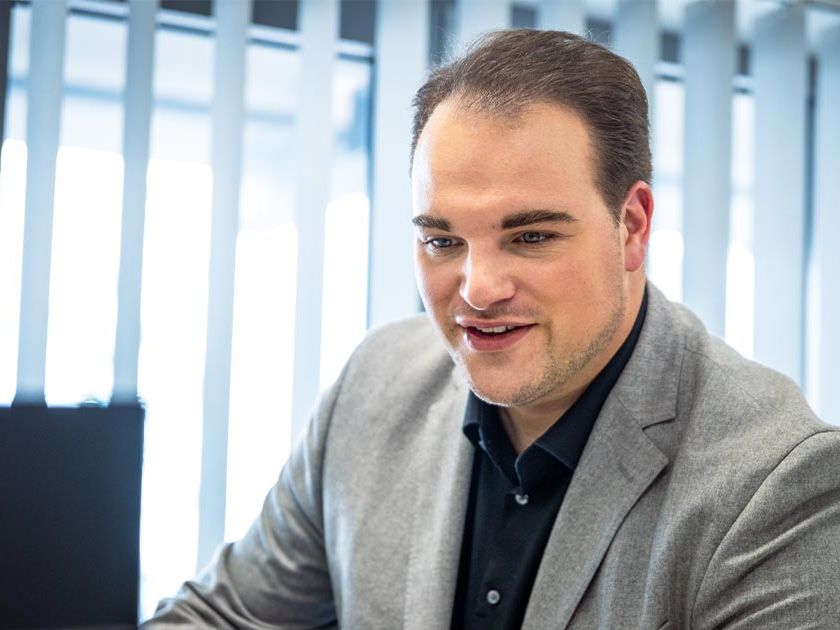 Ein männlicher Mitarbeiter sitzt im Büro vor einem Computerbildschirm und lächelt. Er trägt ein graues Sakko über einem schwarzen Hemd. Im Hintergrund sind vertikale Jalousien an den Fenstern zu sehen.