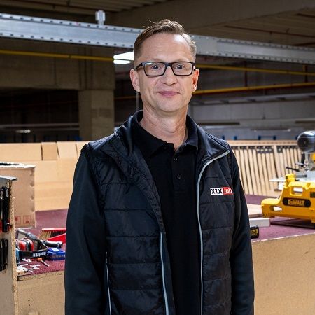Ein Mann mit Brille und schwarzer Weste steht in einer Werkstatt. Im Hintergrund sind eine gelbe elektrische Säge, verschiedene Werkzeuge und Arbeitsmaterialien zu sehen. 