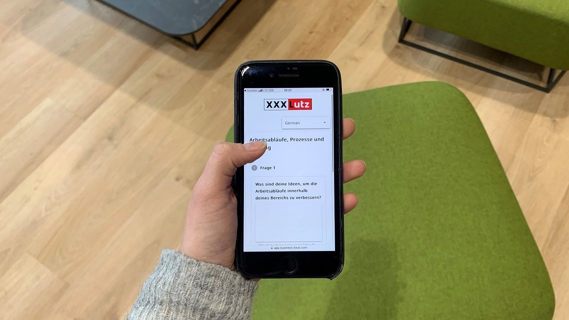 Zu sehen ist ein Auschnitt, wie eine Hand ein Handy in der Hand hält. Auf dem Bildschirm das XXXLutz Logo und ein Teil der Mitarbeiterbefragung "Arbeitsabläufe, Prozesse und..." "Frage 1: Was sind deine Ideen, um die Arbeitsabläufe innerhalb deines Bereichs zu verbessern?"