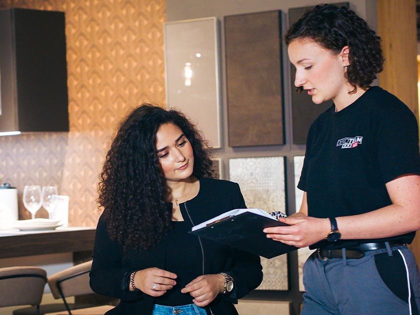 Zwei Frauen stehen in einem modernen Küchenbereich. Die Frau mit lockigem Haar trägt eine schwarze Jacke und Jeans und blickt auf ein Klemmbrett, das von der zweiten Frau gehalten wird. Die zweite Frau trägt eine schwarze Arbeitskleidung und erklärt mit einer Handbewegung etwas auf dem Klemmbrett. Im Hintergrund sind Küchenmöbel, Geschirr und Dekorationselemente wie hängende Lampen und Regale zu sehen.